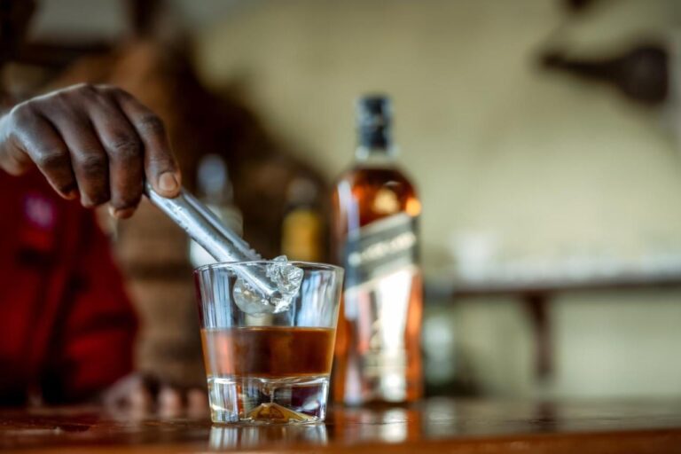 En hånd slipper en isbit i et whiskyglass, på bardisken