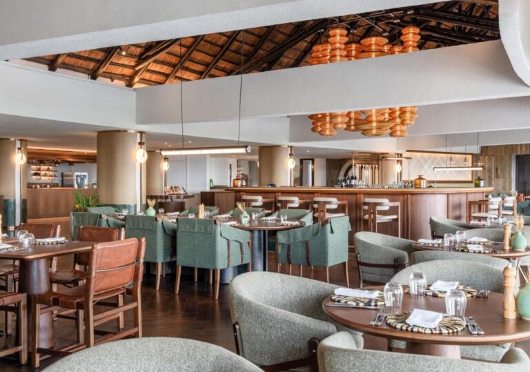 En restaurant med grønne stoler foran en lang bar, under oransje taklamper