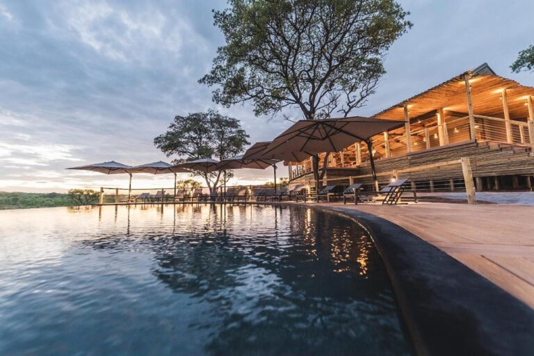 Uendelighetsbassenget på Mdluli Safari Lodge og den opplyste hovedbygningen, under en skyet himmel