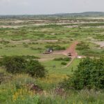 Utsikt fra Observation Hill over en grønn slette med saltflekker, et rødt spor, tre 4x4-biler parkert ved en liten okerfarget bygning og lave åser i det fjerne