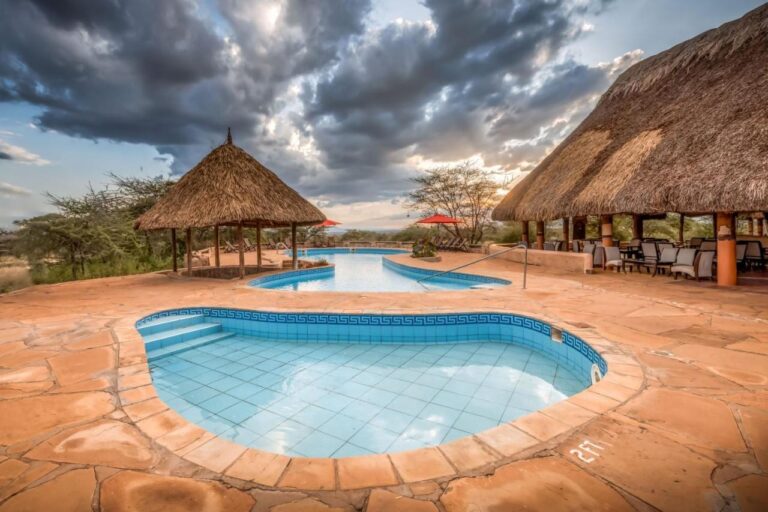 Et lite flislagt basseng i forgrunnen og det store bassenget på Samburu Sopa Lodge, mellom en stråhytte og en terrasse med stråtak